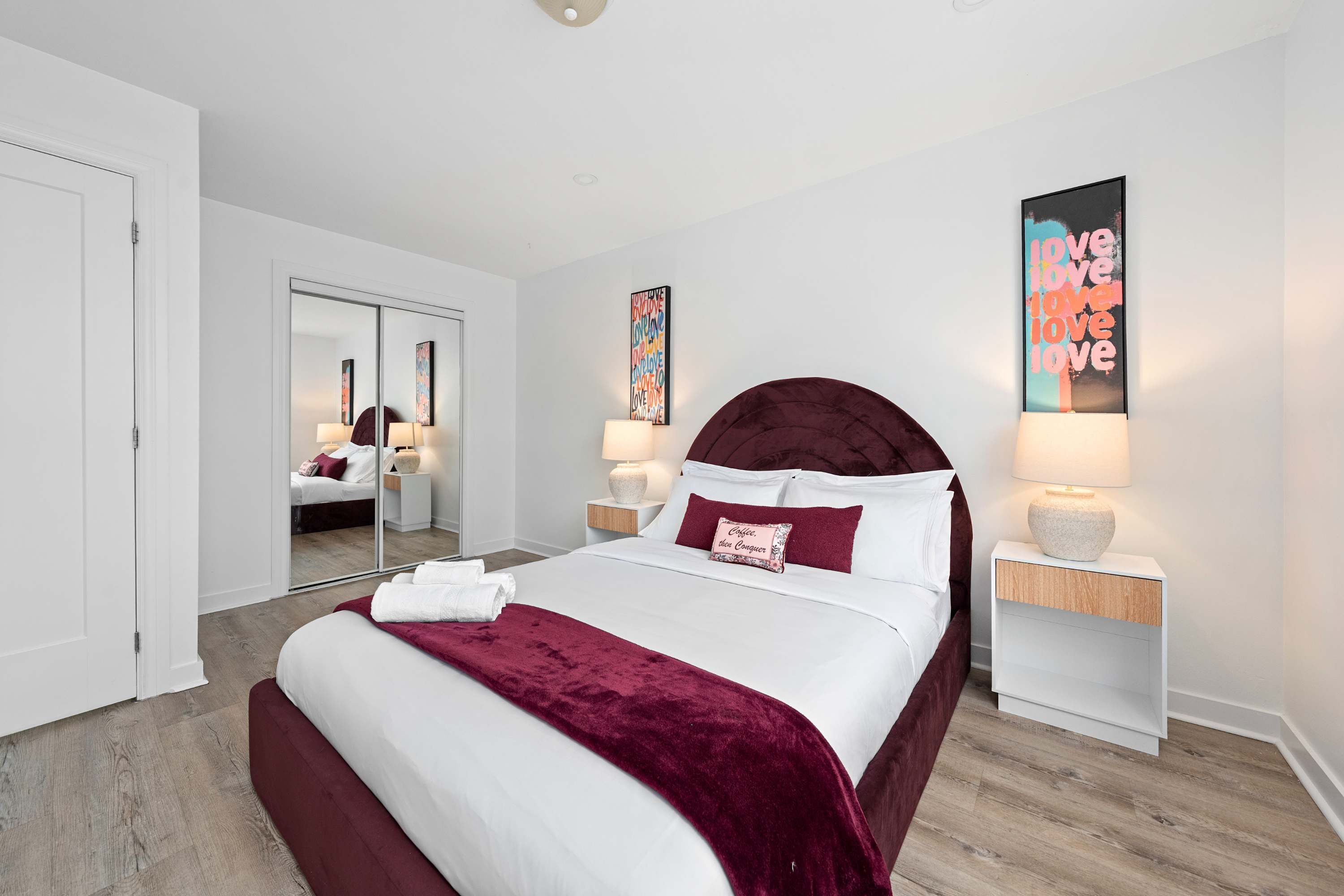Une chambre à coucher moderne avec un grand lit, des tables de chevet avec des lampes, un miroir et des œuvres d’art accrochées aux murs.