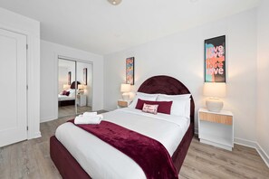 Une chambre à coucher moderne avec un grand lit, un miroir, deux tables de chevet avec des lampes et des œuvres d’art accrochées aux murs.