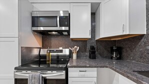 Una cocina moderna con armarios blancos, un microondas de acero inoxidable, una estufa negra y una cafetera.