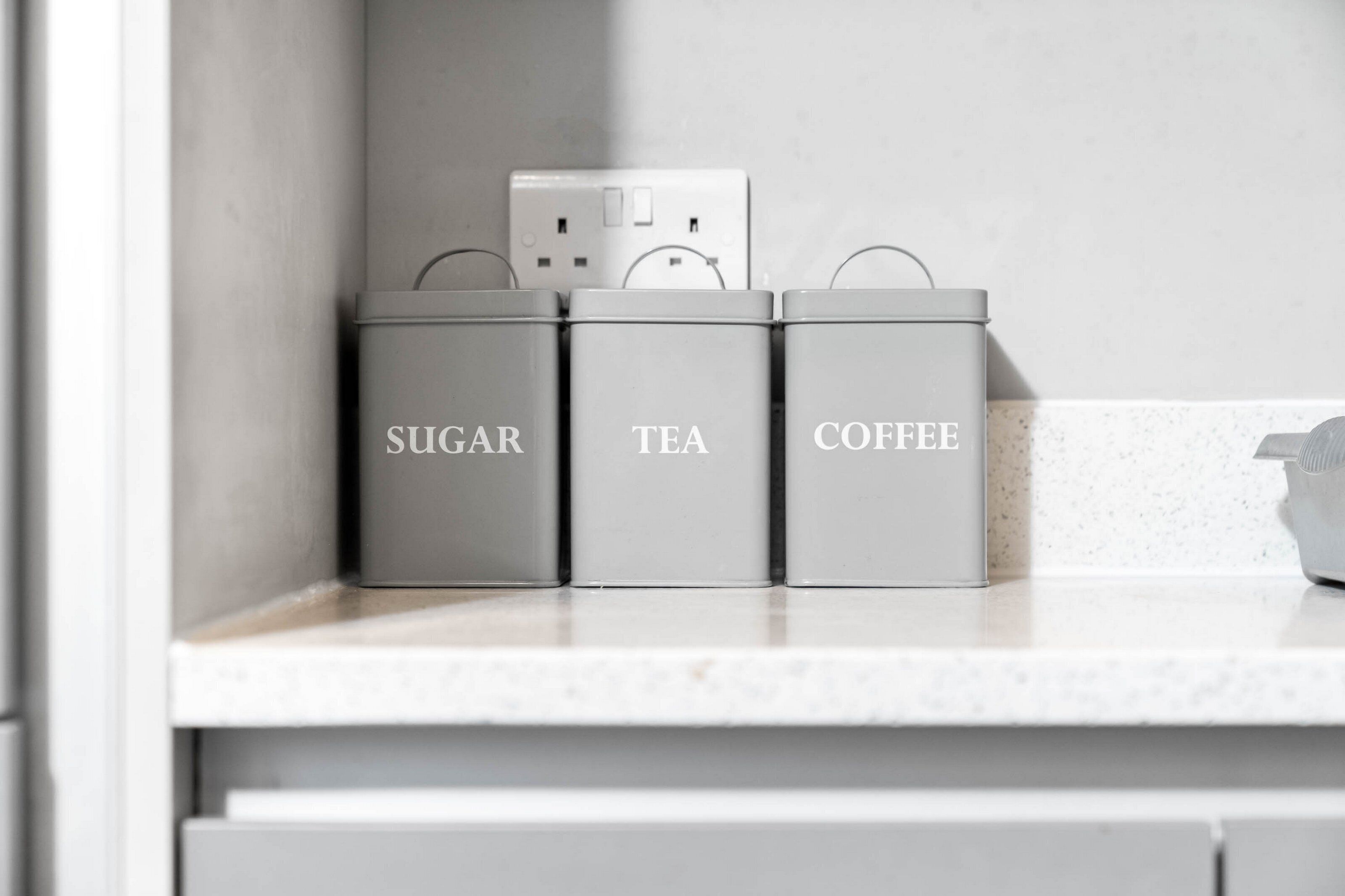 Three gray containers labeled 'SUGAR,' 'TEA,' and 'COFFEE' on a kitchen counter.