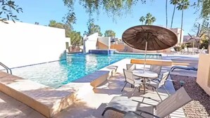 Une piscine entourée de palmiers, avec des chaises longues et un parasol, sous un ciel dégagé.