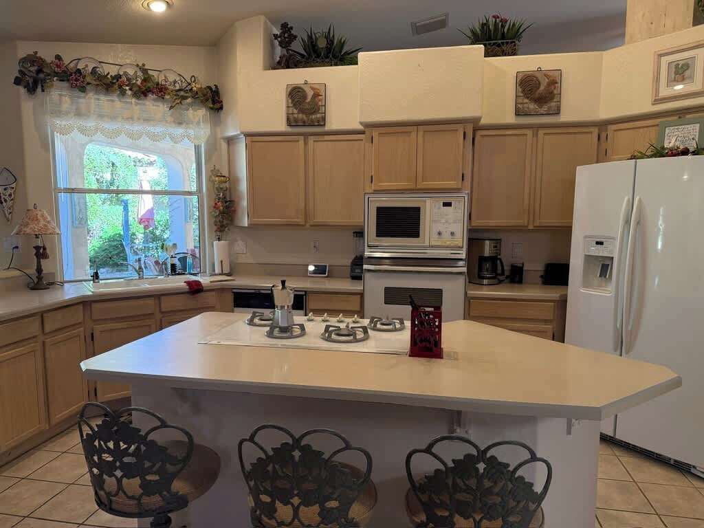A kitchen with wooden cabinets, a central island with chairs, a refrigerator, and a window with a view of greenery.