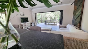 A living room with a sofa, a TV, a coffee table, and a balcony with a view of trees. - Free Standing House in the Middle of Nature (Mbombela)