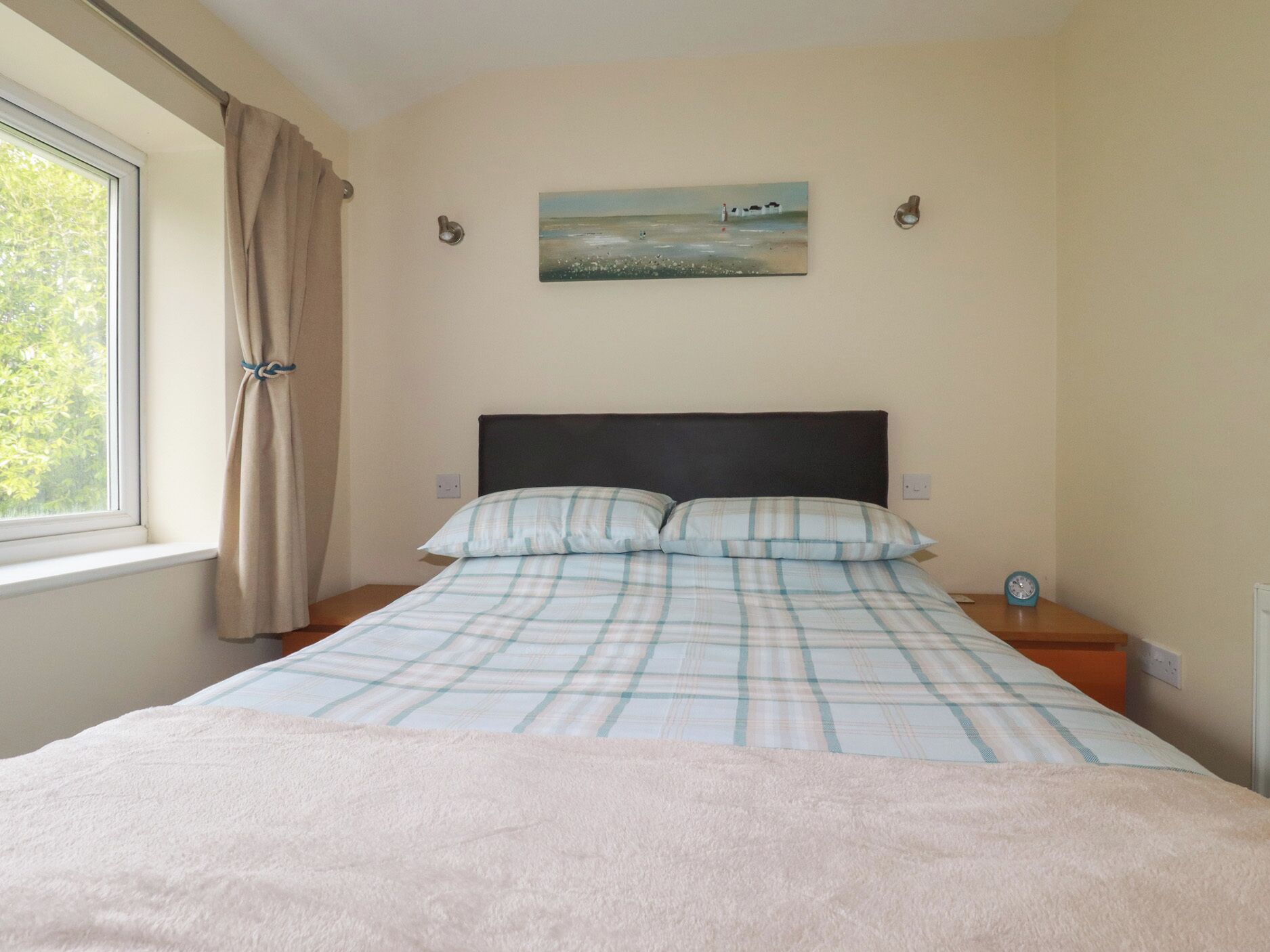 Une chambre à coucher avec un lit, une fenêtre avec des rideaux, un tableau au mur et une table de chevet avec une horloge.