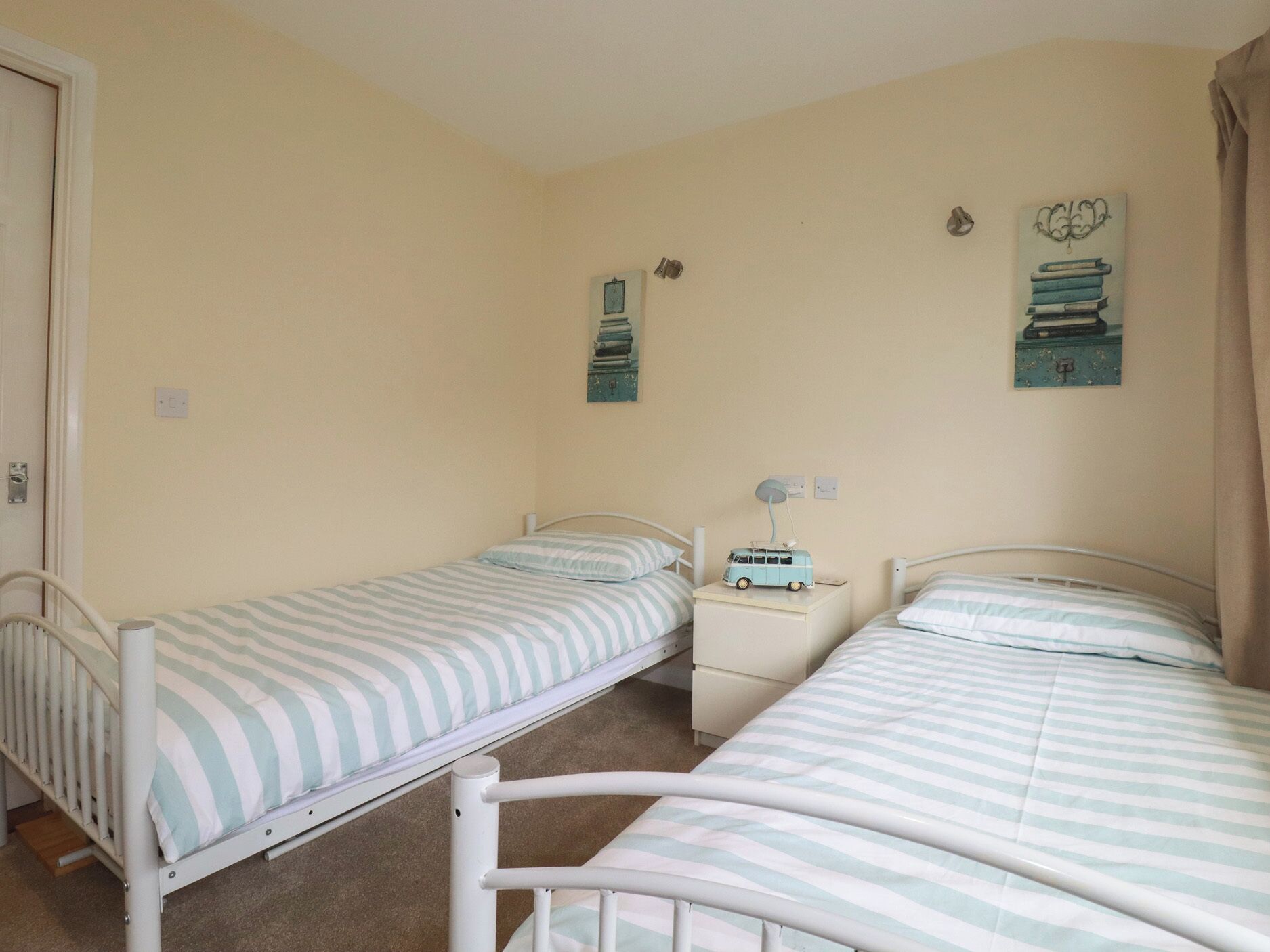 A bedroom with two single beds, a bedside table with a lamp, and a wall-mounted shelf with books and a small toy.