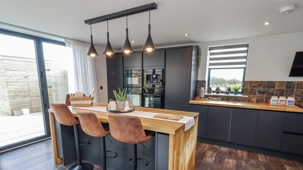 A modern kitchen with a wooden island, bar stools, pendant lighting, and a view of the outdoors. - The Joiners Cottage (Morpeth)