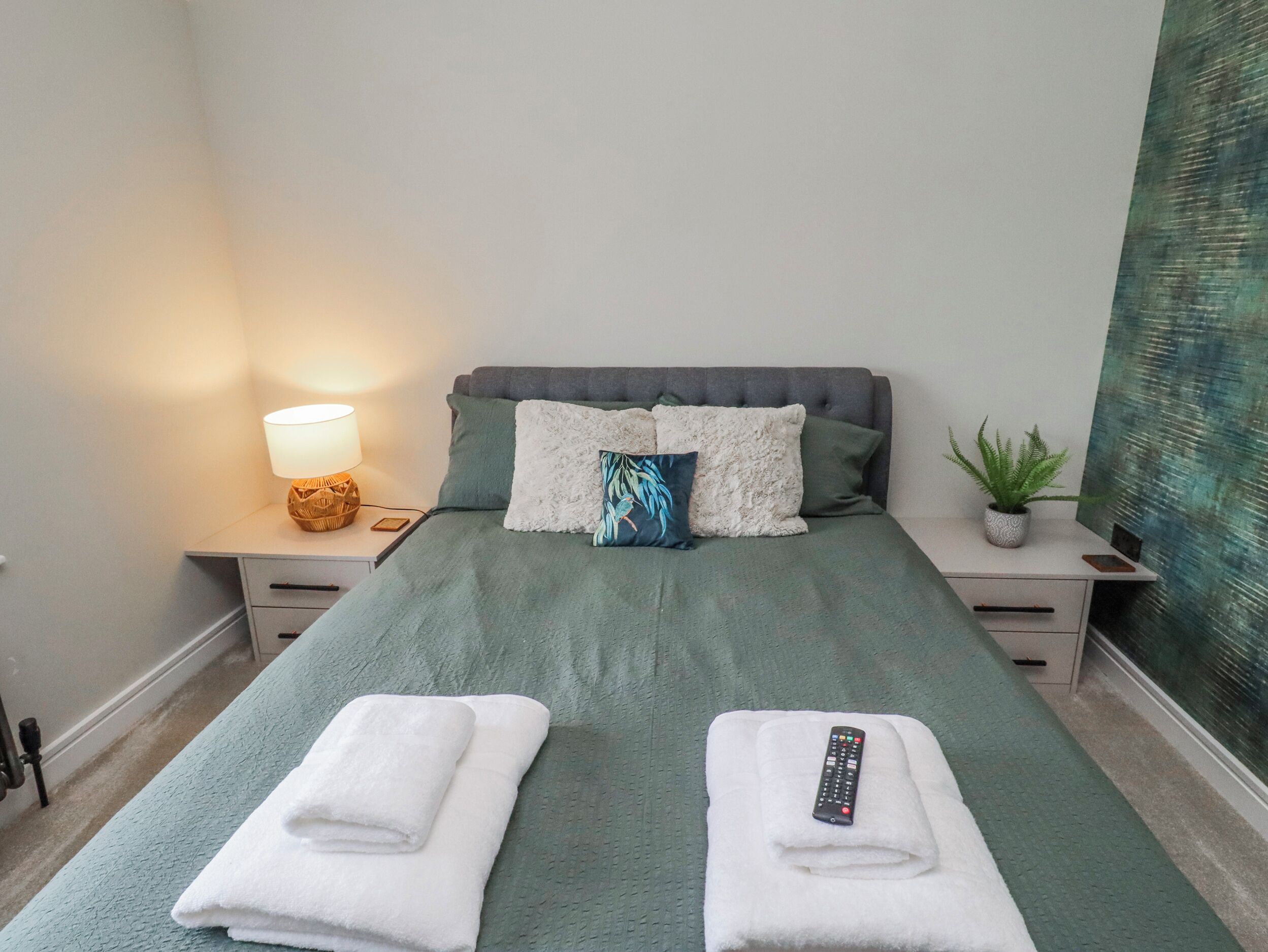 A neatly made bed with a green bedspread, two white towels, and a remote control on the bed. There is a lamp on the nightstand beside the bed, a potted plant on the nightstand, and a wall with a textured design.