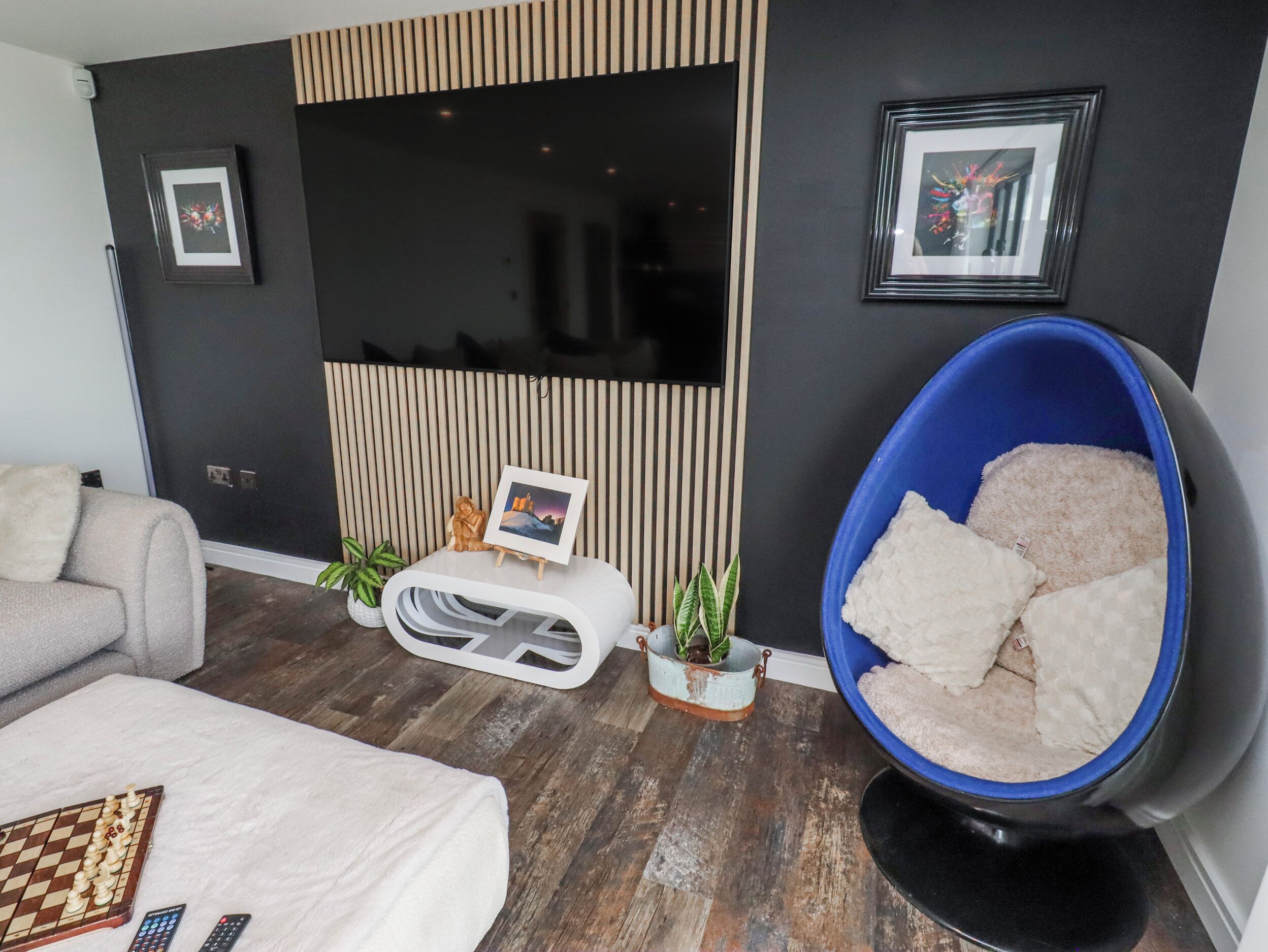 A modern living room with a flat-screen TV, a blue bean bag chair, a sofa, a chessboard, and a small table with a picture frame and plants.