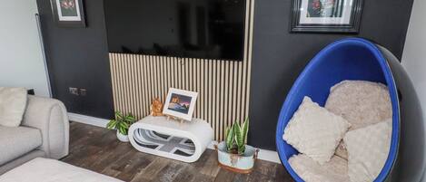 A modern living room with a flat-screen TV, a blue bean bag chair, a sofa, a chessboard, and a small table with a picture frame and plants.