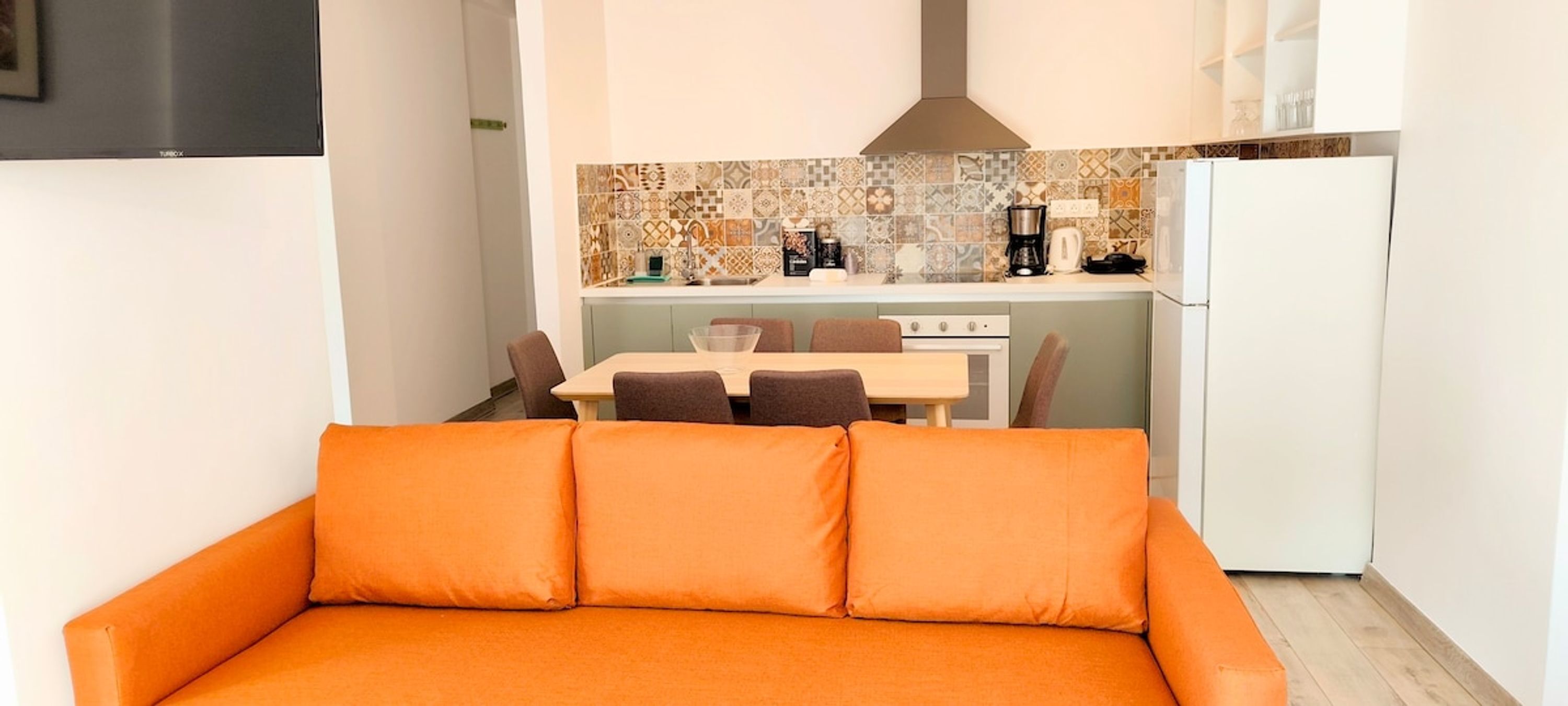 A compact living space with a sofa, a dining table with chairs, a kitchenette, and a white refrigerator.