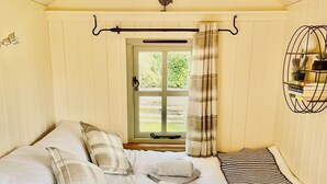 Een slaapkamer met een bed, kussens, een raam met gordijnen, een radiator, en een wandplank met boeken en planten.