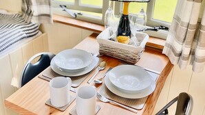 Een eettafel gedekt met borden, bekers en bestek, een fles wijn en een mand met kranten.