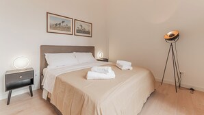 A neatly made bed with white linens, a bedside table with a lamp, and a floor lamp in a room with light wooden flooring and plain walls. - Casa Martini - Vatican City (Rome)