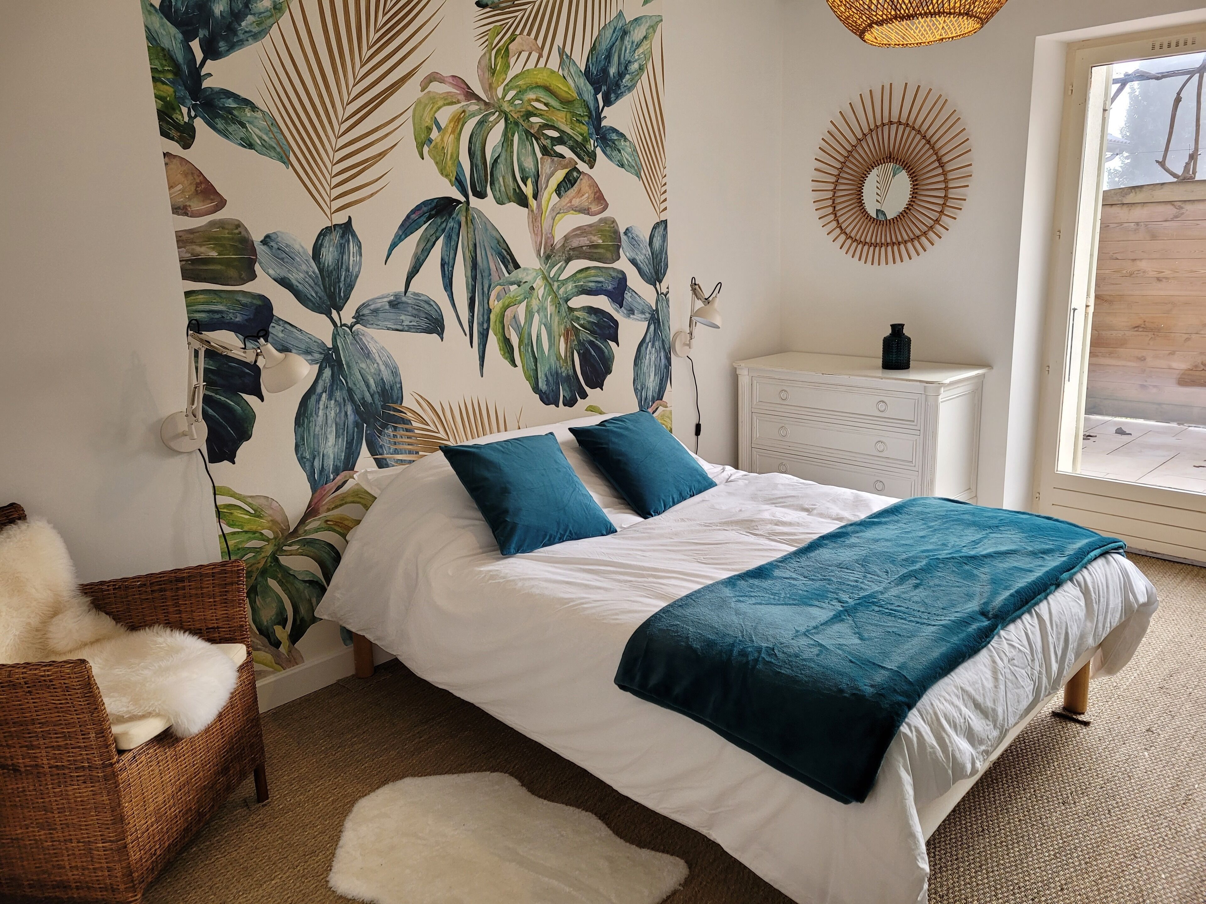 Una camera da letto con un letto, una cassettiera bianca e una parete con carta da parati a motivi tropicali.