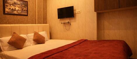 A bed with a red blanket, two pillows, a mounted TV, and a wall-mounted air vent.