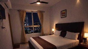 A hotel room with a bed, a desk, a chair, a ceiling fan, a window with curtains, and a night view of the city. - Glitz Nand -Nr Patna Railway Stn (Patna)