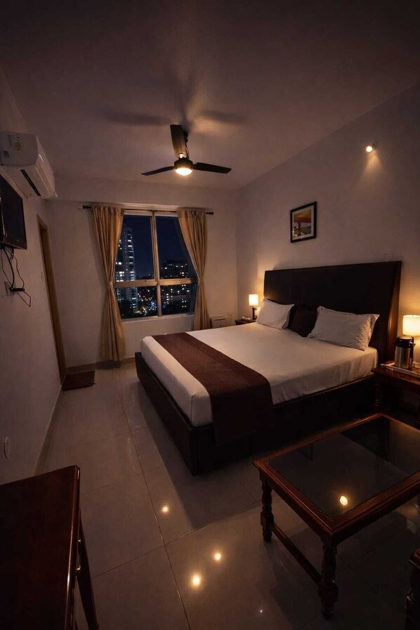 A hotel room with a bed, a desk, a chair, a ceiling fan, a window with curtains, and a night view of the city. - Glitz Nand -Nr Patna Railway Stn (Patna)