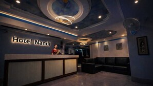 A hotel reception area with a reception desk, a receptionist, a seating area with a sofa, and a ceiling with circular light fixtures. - Glitz Nand -Nr Patna Railway Stn (Patna)