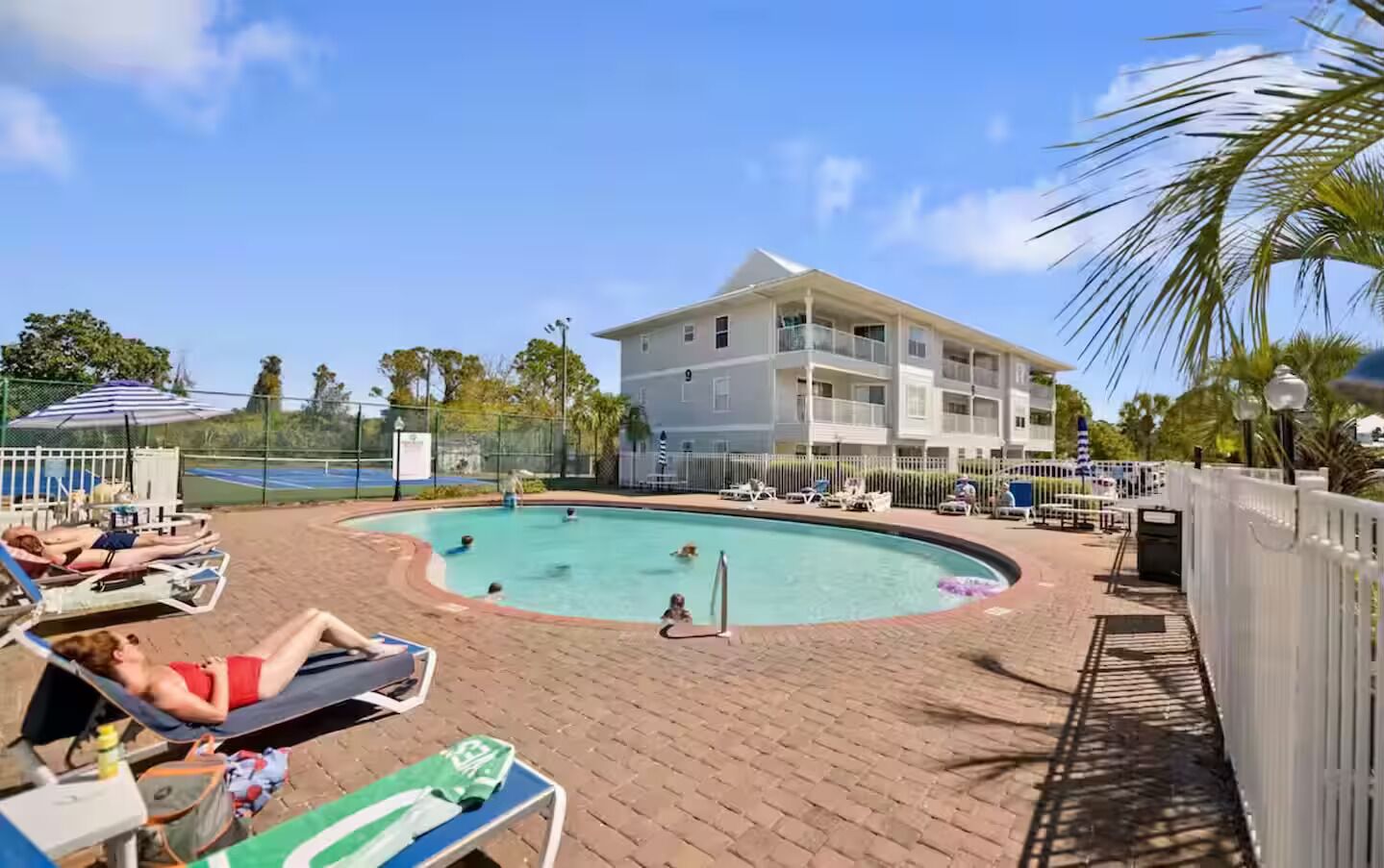 A pool area with lounge chairs, a building with balconies, and a fenced tennis court.