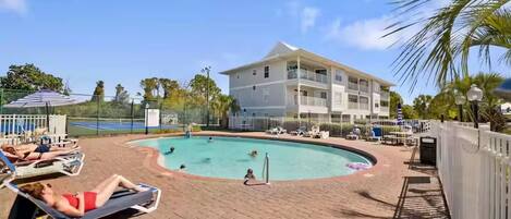 A pool area with lounge chairs, a building with balconies, and a fenced tennis court.