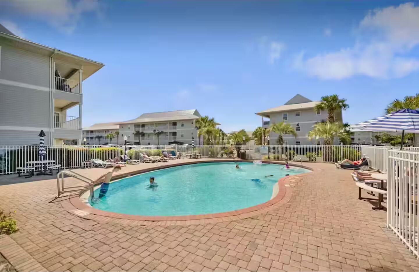 A pool area with lounge chairs and umbrellas, surrounded by a fence and buildings.
