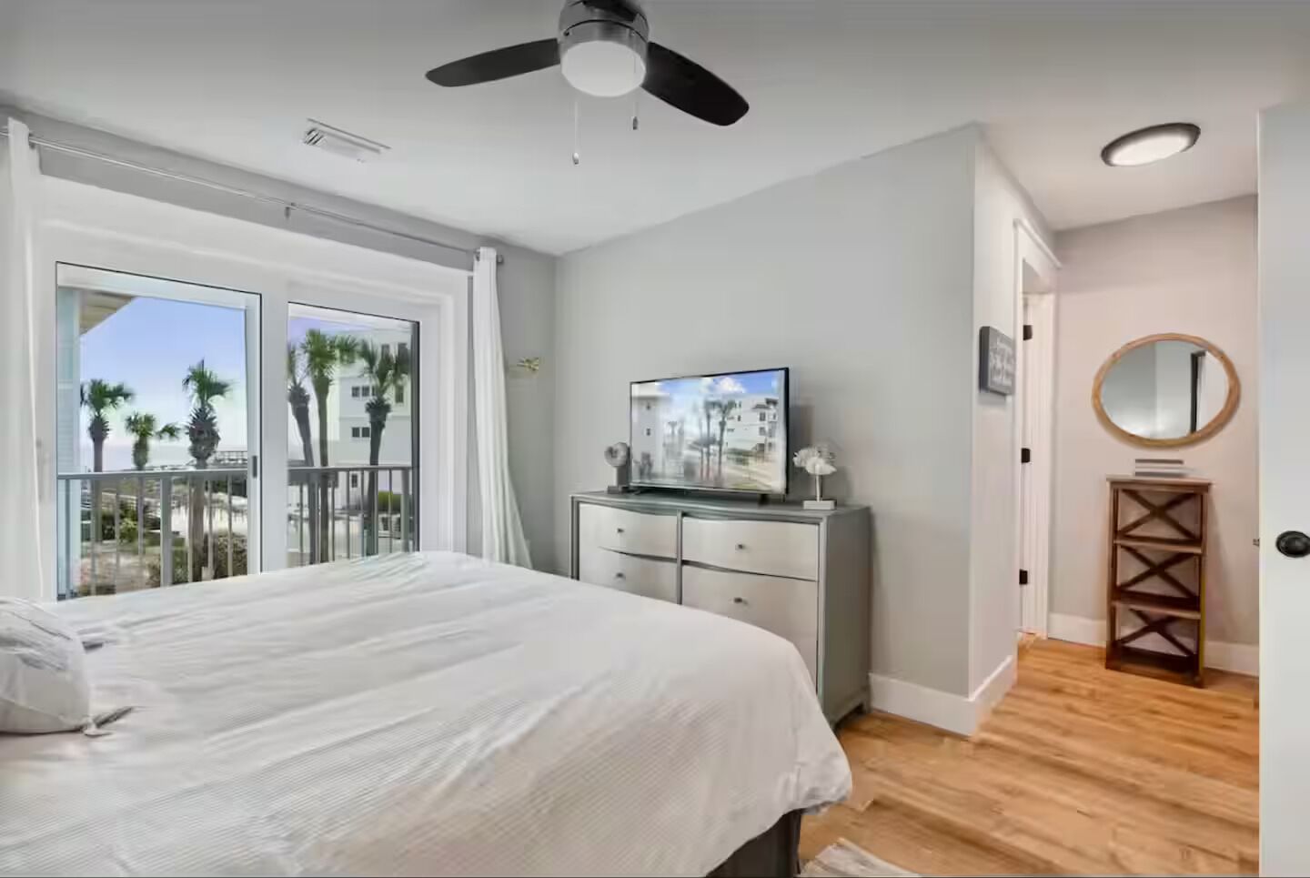 A bedroom with a bed, a dresser, a television, and a balcony with a view of palm trees.