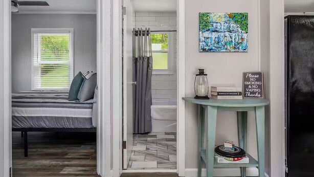 A bedroom with a bed, a window with blinds, a shower area, a small table with a sign, and a painting on the wall. - New-Sunfish Cottage - Walk to Hidden Harbor Marina (Smithville)