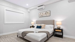Ein modernes Schlafzimmer mit einem großen Bett, zwei Nachttischen mit Lampen, einer Wand-Klimaanlage und einem großen Fenster.