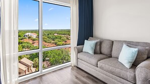 A living room with a large window, a grey sofa, and a view of a residential area.