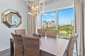 A dining area with a large table, wicker chairs, a chandelier, and a city view through large windows. - 🌊🌴Gulf Views | Walk to the Beach | Lagoon Pool | Luau II 6832 (Miramar Beach)