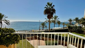 Een balkon met een witte tafel en stoelen, met uitzicht op een strand met palmbomen en de oceaan.