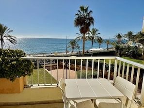 A balcony with a white table and chairs, overlooking a beach with palm trees and the ocean. - 0. Diana 101 (Marbella)