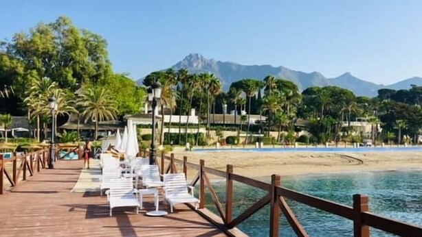 Een houten steiger met witte lounge stoelen en parasols, die naar een strand leidt met palmbomen en bergen op de achtergrond.