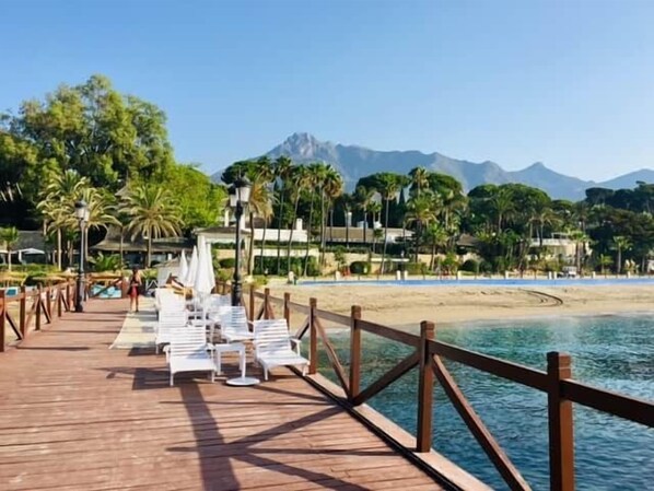 A wooden pier with white lounge chairs and umbrellas, leading to a beach with palm trees and mountains in the background. - 0. Diana 101 (Marbella)