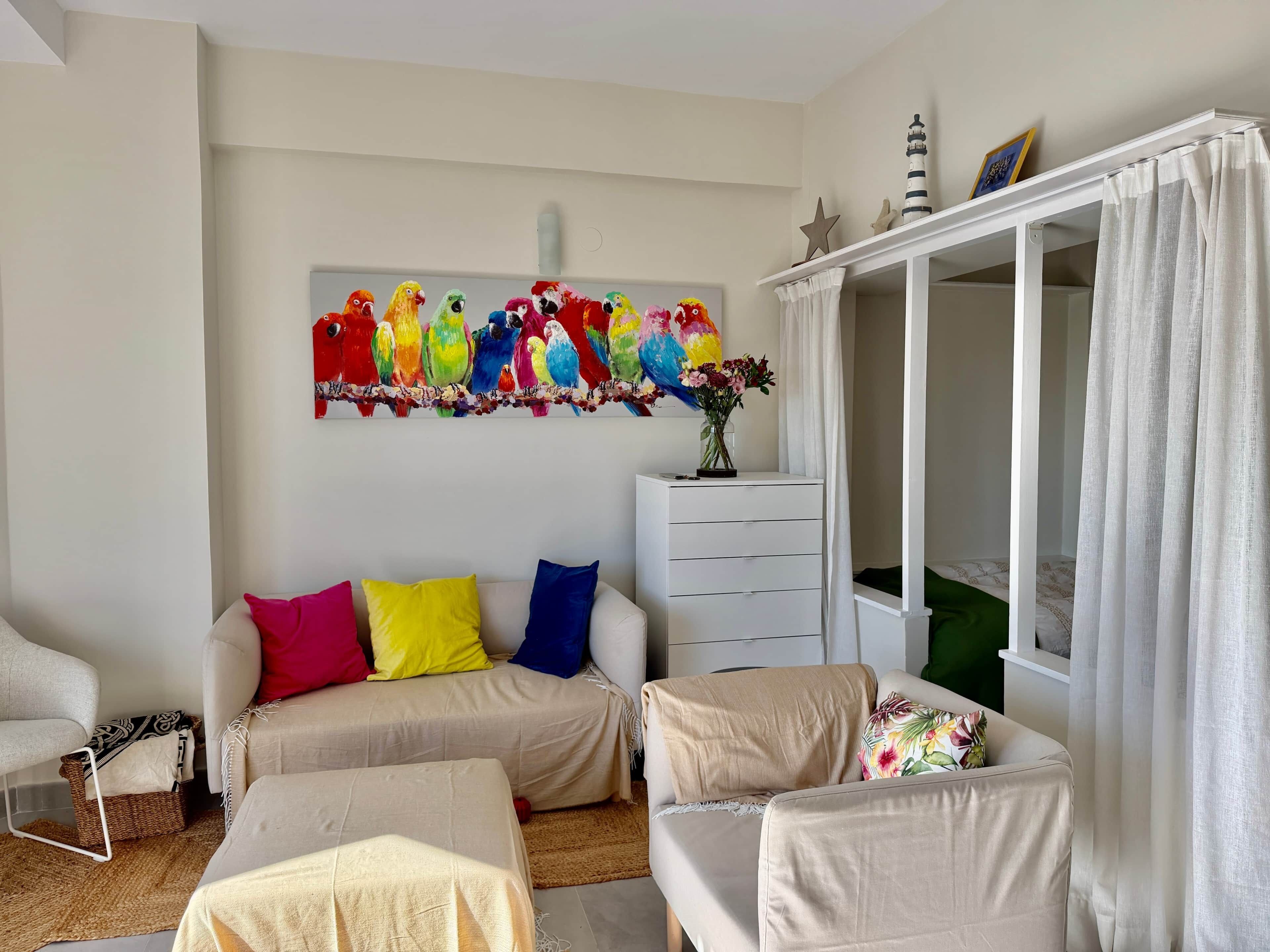 Een woonkamer met een zithoek, een wit kastje, een schilderij van kleurrijke vogels, en een raam met gordijnen.