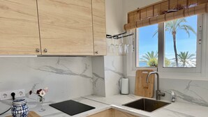 A kitchen with marble countertops, wooden cabinets, a black cutting board, a wooden cutting board, a stainless steel sink, a window with a view of palm trees, and a bamboo blind. - 0. Skol Marbella 111A (Marbella)