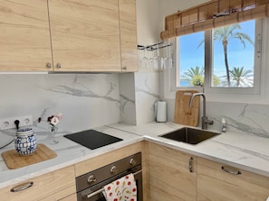 A kitchen with marble countertops, wooden cabinets, a black cutting board, a wooden cutting board, a stainless steel sink, a window with a view of palm trees, and a bamboo blind. - 0. Skol Marbella 111A (Marbella)