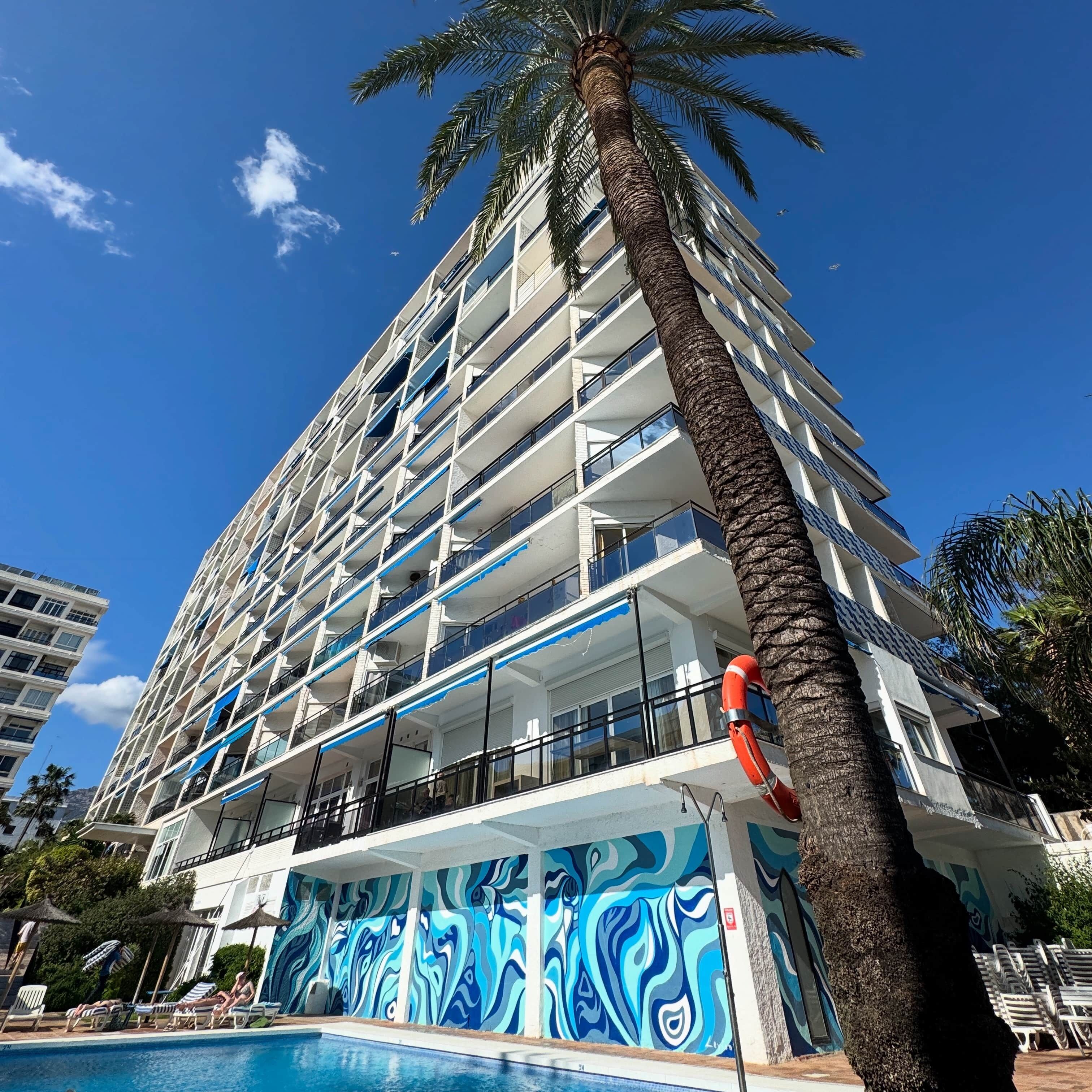 A multi-story hotel with balconies, a swimming pool, and palm trees.