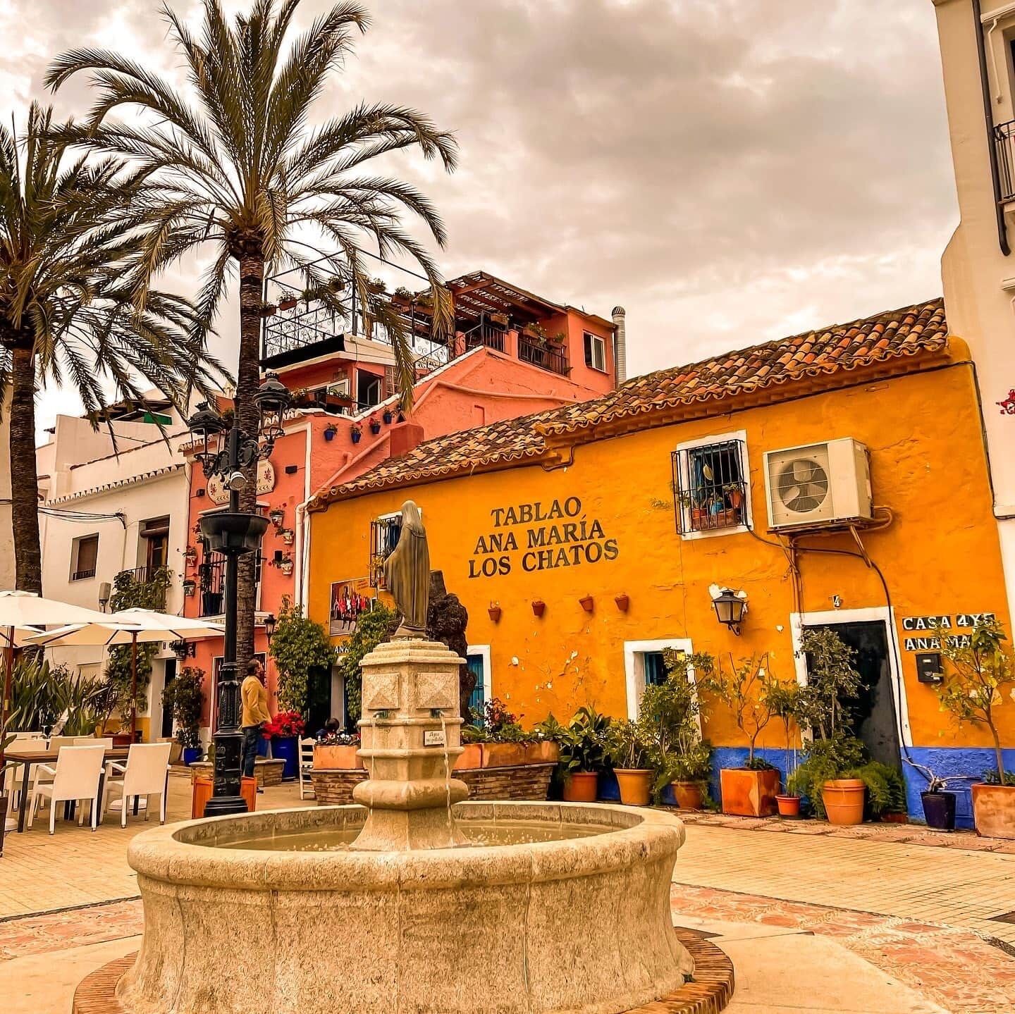 A courtyard with a fountain, palm trees, and a building with 'TABLAO ANA MARIA LOS CHATOS' written on it.