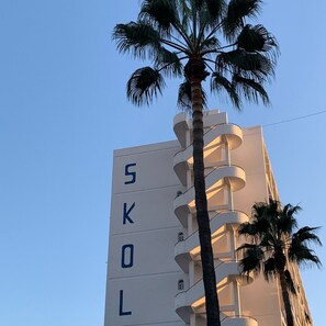 Ein Gebäude mit vertikal angeordneten Buchstaben 'SKOL' an der Fassade, umgeben von Palmen.