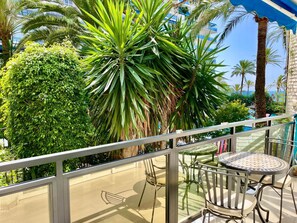 Ein Balkon mit Glastrennwand, einem runden Tisch und Stühlen, mit Blick auf einen üppigen Garten mit verschiedenen Pflanzen und einem Gebäude im Hintergrund.