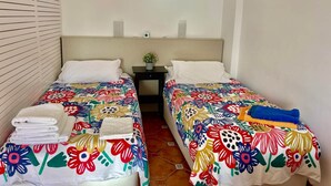 Una habitación de hotel con dos camas, cada una con una colcha floral de colores, una mesita de noche con una planta y un estante en la pared.