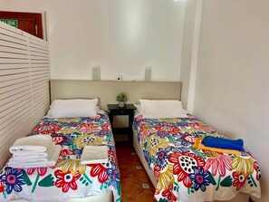 Ein Hotelzimmer mit zwei Betten, jeweils mit farbenfroher Blumenbettdecke, einem kleinen Nachttisch mit Pflanze und einem an der Wand befestigten Regal.