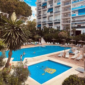 Ein Schwimmbecken mit einem Cartoon-Design, umgeben von Liegestühlen und Sonnenschirmen, mit einem mehrstöckigen Gebäude im Hintergrund.