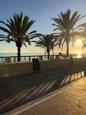 Eine Küstenpromenade mit Palmen, blauem Himmel und der Sonne, die über dem Meer aufgeht oder untergeht.