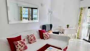 Ein modernes Wohnzimmer mit einem weißen Sofa, roten und gemusterten Kissen, einem weißen Couchtisch, einem Essbereich mit Tisch und Stühlen sowie einem großen Fenster mit Ausblick.