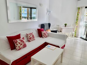 Ein modernes Wohnzimmer mit einem weißen Sofa, roten und gemusterten Kissen, einem weißen Couchtisch, einem Essbereich mit Tisch und Stühlen sowie einem großen Fenster mit Ausblick.