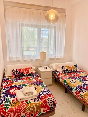 Ein Zimmer mit zwei Betten, einem Nachttisch, einer Lampe und einem Fenster mit durchsichtigen Vorhängen.