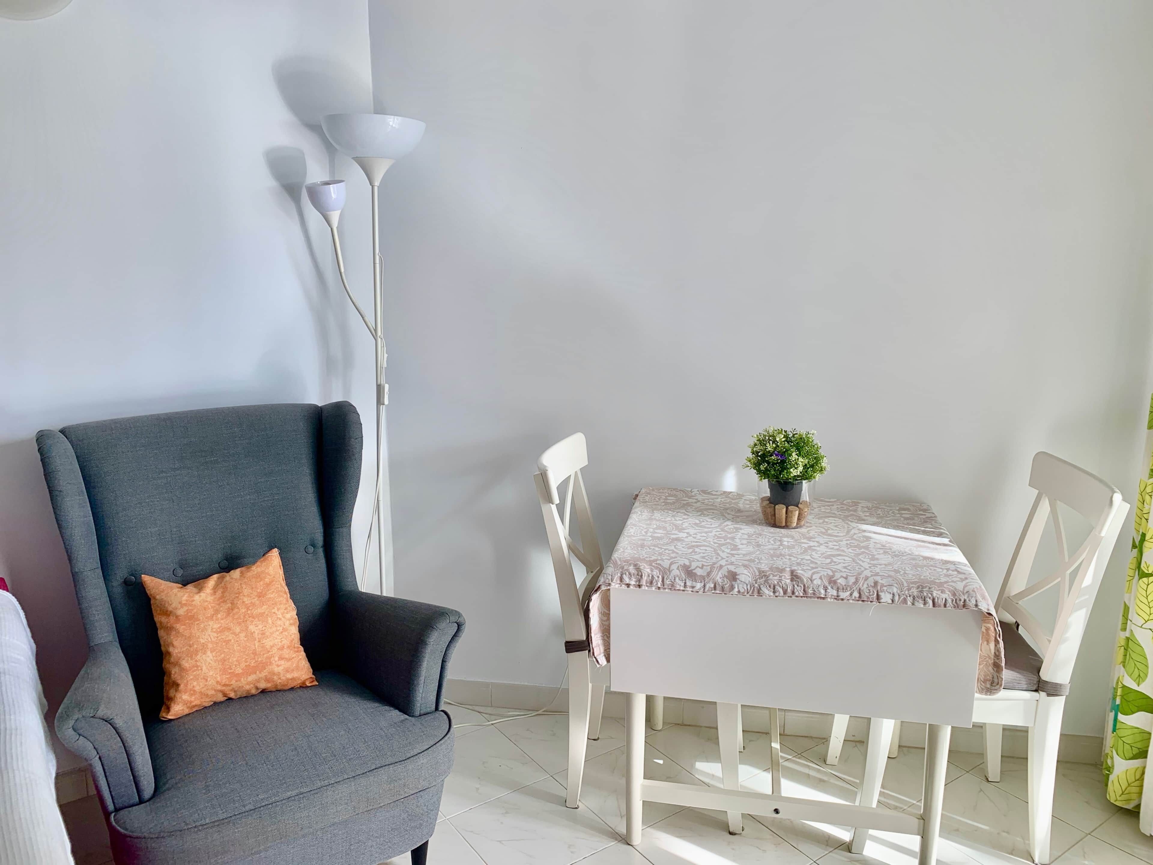 Una habitación con un sillón gris, una mesa blanca con mantel estampado, una pequeña planta en maceta y una lámpara de pie blanca.