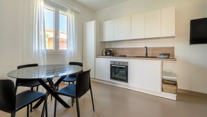 Una cocina moderna con armarios blancos, horno empotrado y comedor con mesa de vidrio y sillas negras.